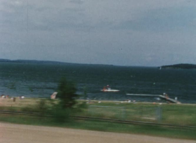 People on a beach and boats in the lake as seen from a car driving
