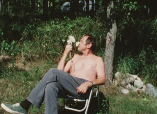 Shirtless man sitting on a folding chair smells a flower while waving caught on vintage film