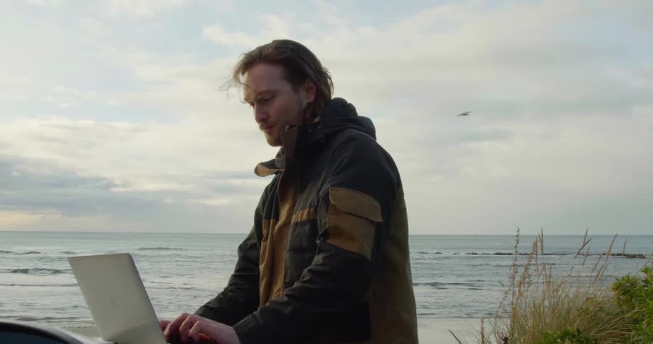 A man is parked along the coast and standing outside typing on his laptop computer on the hood of his car