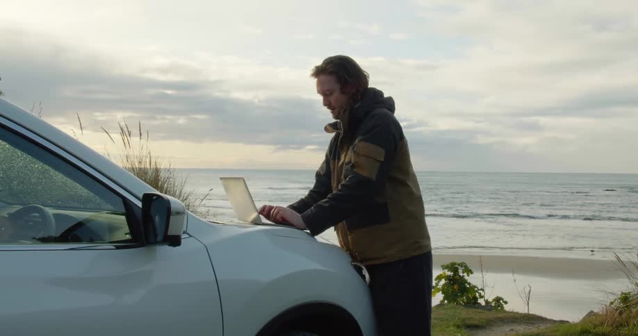 A man is parked along the coast and standing outside typing on his laptop computer on the hood of his car