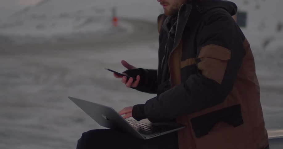 Man sits outdoors near the mountains in the winter and looks at his smart phone and types on a laptop