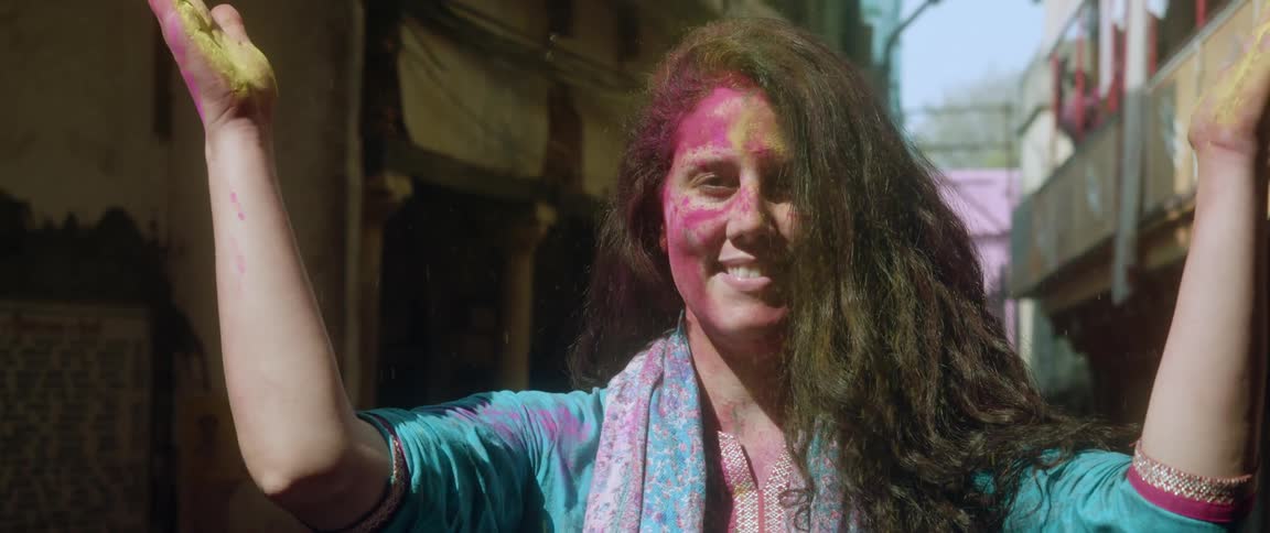 A teenage girl with paint on her face and hands stands between buildings and claps so the dye on her hands flies in the air