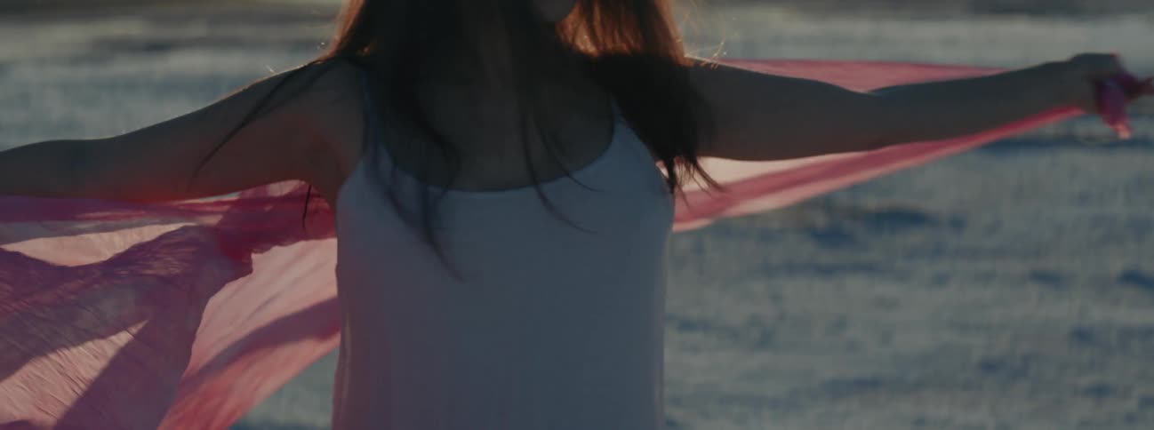 A woman spinning around on a beach with a pink shall around her shoulders as the sun shines over the horizon