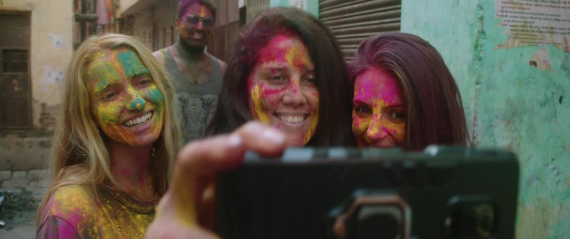 A group of women wearing face paint pose for a smartphone photo while a man tries to sneak into the photo in the background