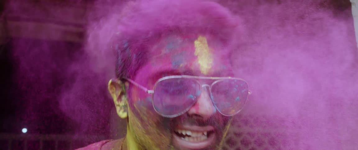 A man with his face hair beard and sunglasses covered in powdered dye shakes his head spreading the dye around him