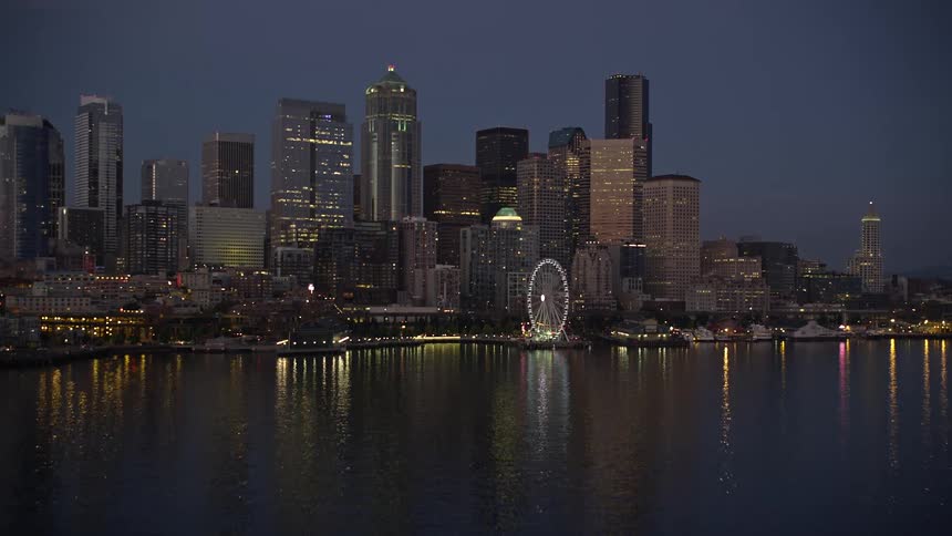 Low flying aerial footage captures the clear twilight skyline of Downtown Seattle Washington as the camera approaches the city revealing high rise buildings the iconic ferris wheel and the shoreline reflecting in the water near docks