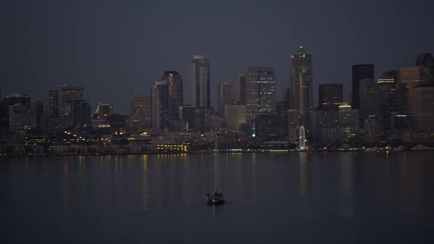 A sailboat sails in the harbor of Seattle towards the Downtown skyline at twilight showcasing the city's high rise towers and waterfront recreation