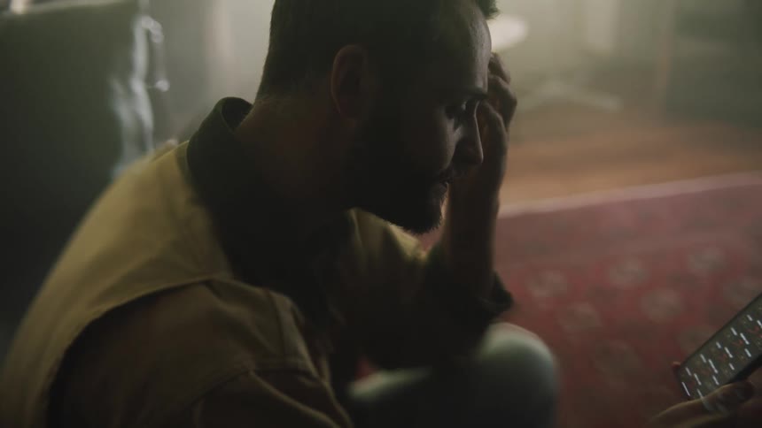 A close up of a man sitting in his living room looking at his cellphone contemplatively