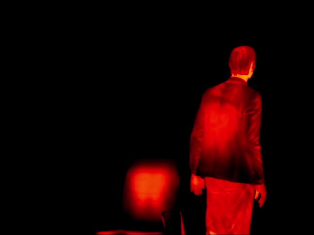 Male image colored in reds and yellows contrasts with black surroundings as he gets up from chair and walks away