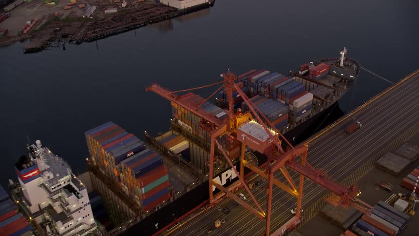 Flying over cargo ships laden with containers docked beneath cranes at the Port of Seattle Washington at sunset