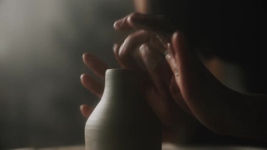 Female potter shapes clay vessel turning on pottery wheel in dimly lit area