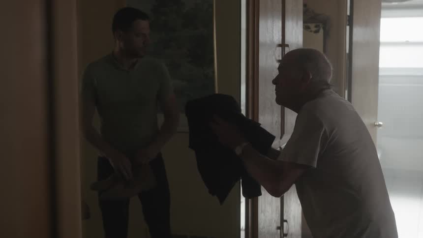 An elderly man holding a coat argues in a hallway with a younger man holding a pair of shoes throwing the coat and shouting at the younger man when he walks away