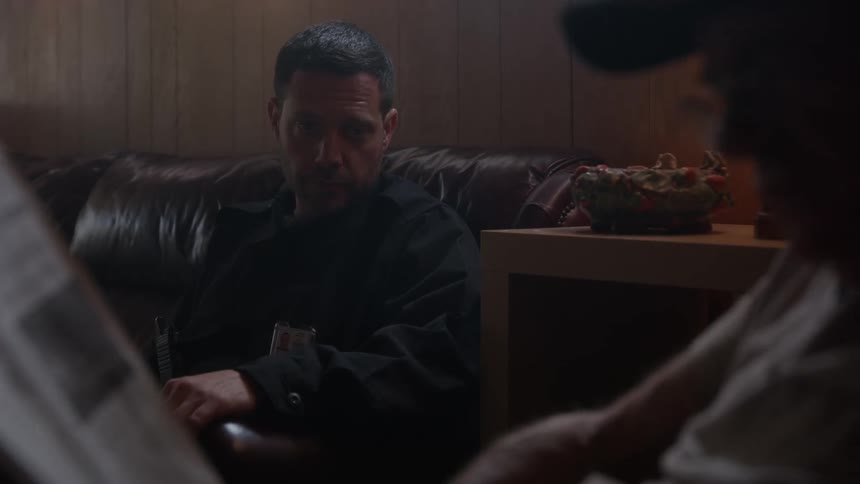 A man sits on a couch and sips from a glass of orange juice then tries making conversation with the elderly man sitting in a chair in front of him reading a newspaper who reacts badly making the man get up and leave