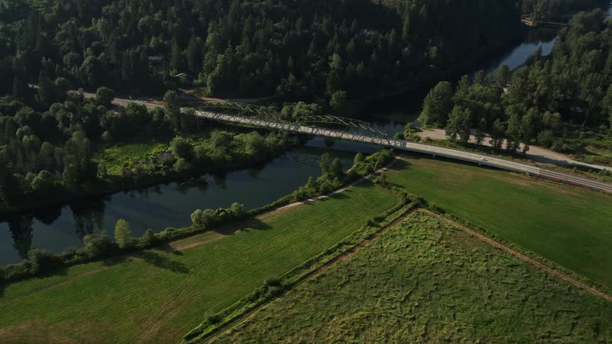 Aerial footage tracks over a green field and approaches a small bridge spanning the Snoqualmie River in Carnation Washington during the daytime