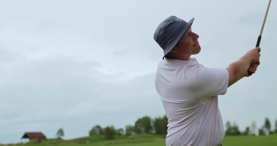 A man swings a golf club and licks his lips as he follows the ball after the strike