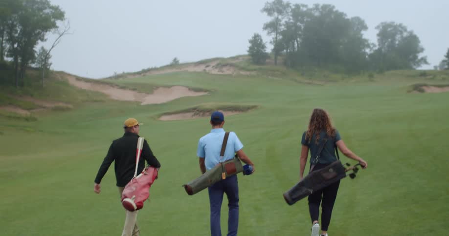 Three golfers carry their golf clubs while walking across golf course on misty day