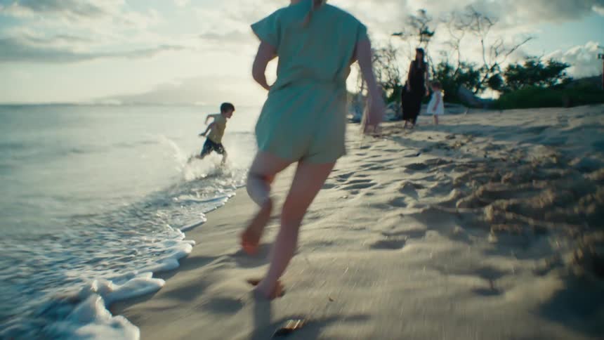 A girl chases her brother toward the ocean as their family watches