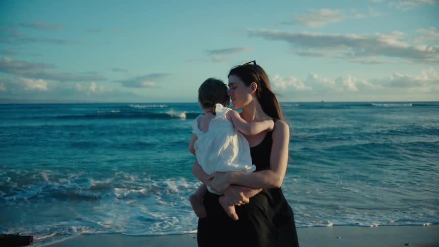 A woman holds her baby while standing on the beach along the ocean