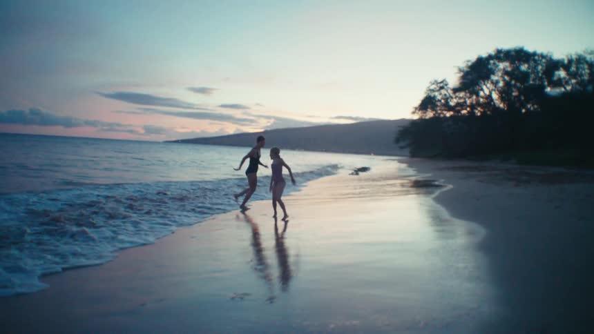 A woman chases a young girl on a beach at waters edge as they both smile and laugh and the sun sets