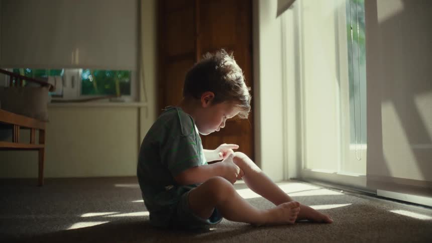 A toddler sitting on the floor inside trying to get something off his knee