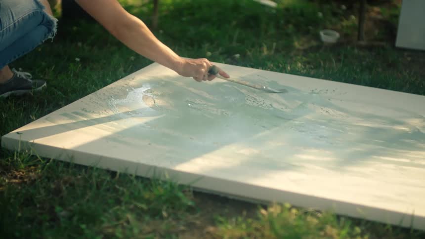 A man uses a small brush to paint a canvas on a grassy ground