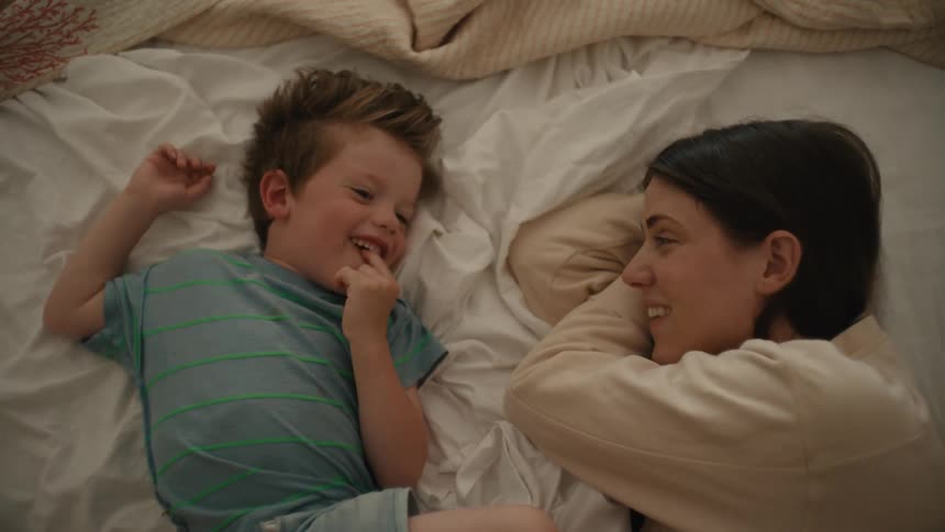 A boy laughs while lying in bed with his mom