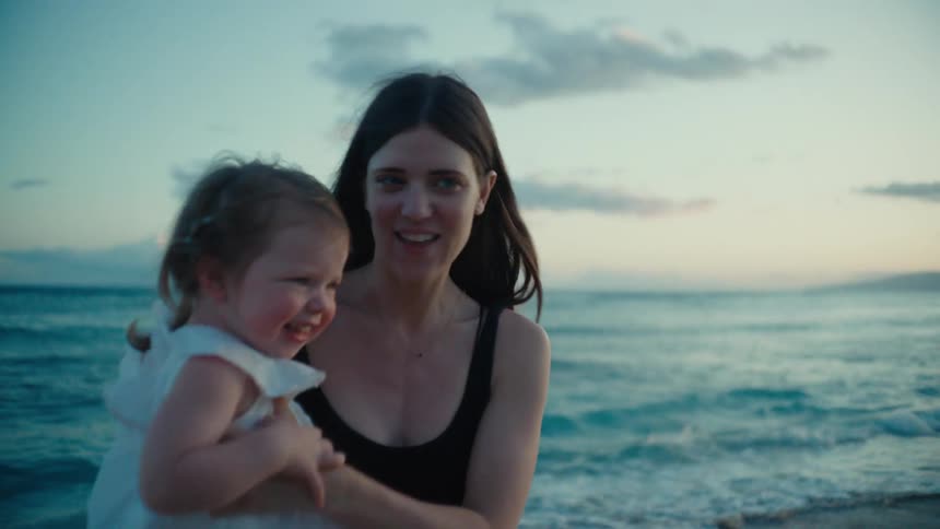 A woman on a beach is holding a baby girl in her laugh and they both smile and laugh as the woman spins and dances with the baby