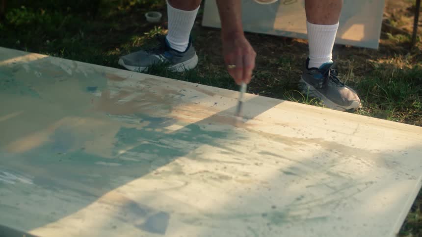 Painter with white socks and sneakers moves brush across the canvas laying on green lawn