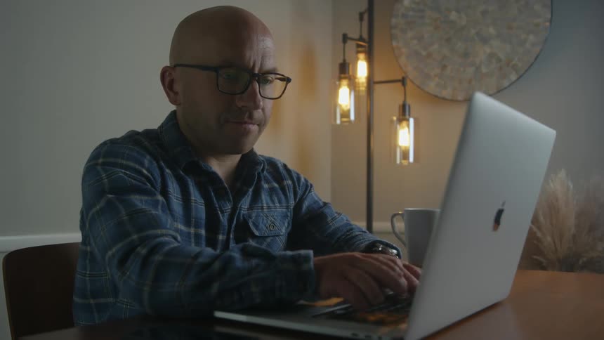 Bald mand with glasses and blue shirt sits at desk working on silver macbook laptop in room