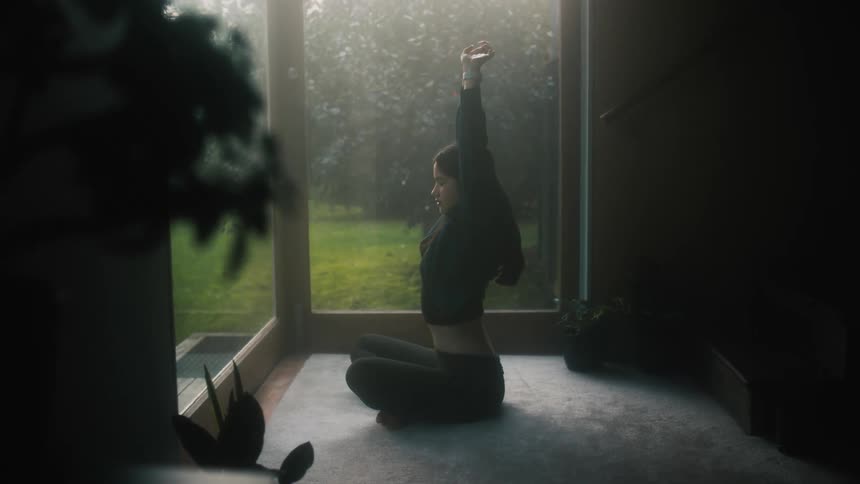 Woman sits crosslegged on carpet near floor to ceiling glasses and stretches