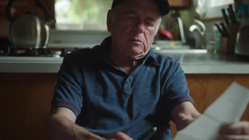 Elderly man sits at table noting down bills while budgeting