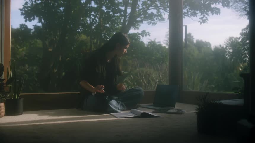Woman sits on floor near window taking notes from laptop