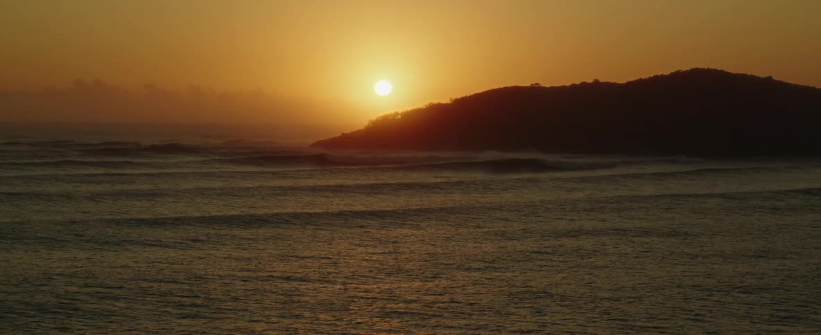 Ocean waves roll into a hillside shore under a warm sunset