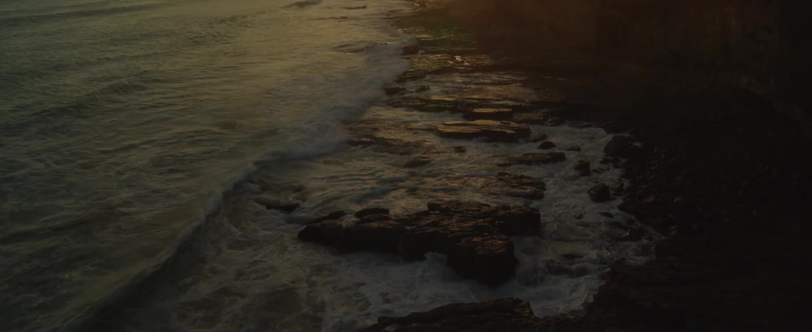Waves roll over rocks along the coast and roll in to the beach under the light of the setting sun