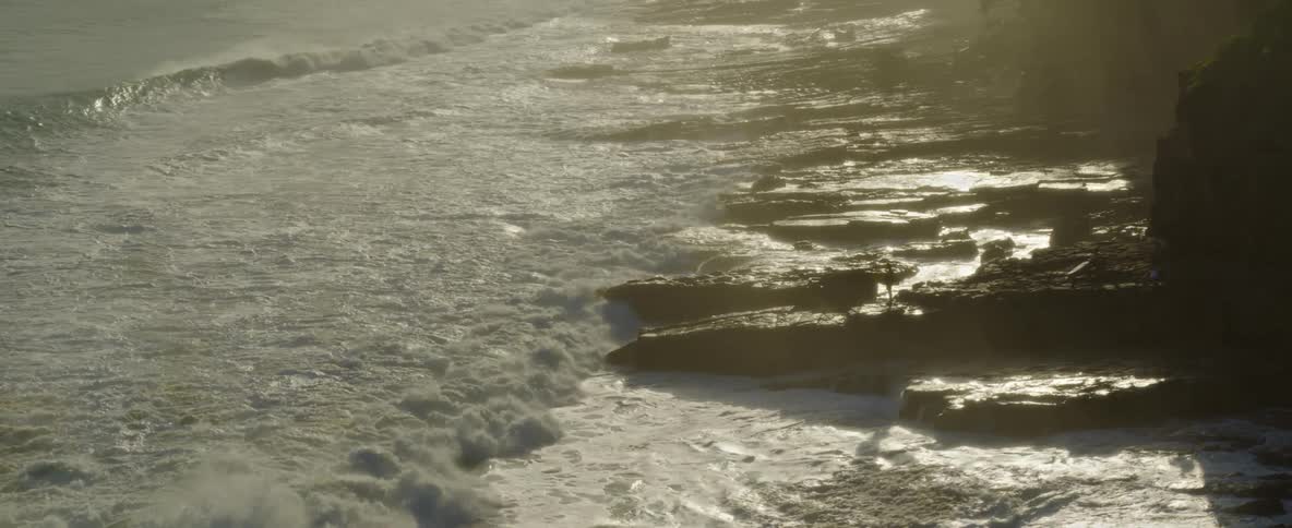 An aerial of the ocean meeting rocks on the beach as people stands on the rocks while waves crash up