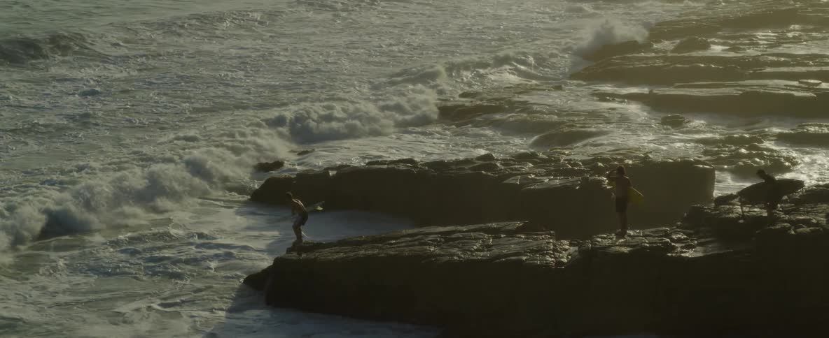 Drone view of waves crashing against the rocky coast as the silhouette of people cascade shadows off the rocks