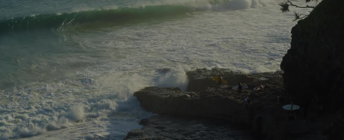An aerial shot of two surfers standing on a rocky outcropping as waves crash against the shore