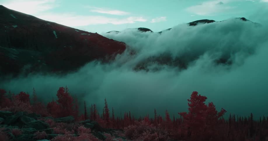 Dense fog flows over color filter red tinted vegetation near mountain range on cloudy day