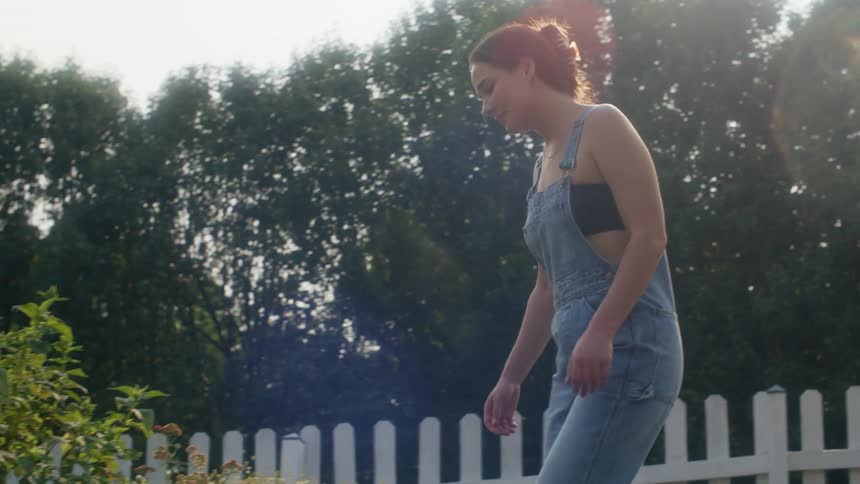 Woman walks across yard and crouches to examine plants in garden bed