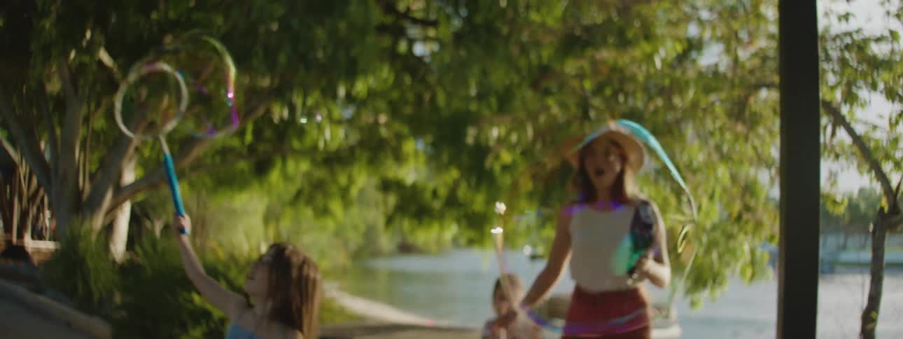 A mother and her daughters playing with bubbles outside near a lake behind them