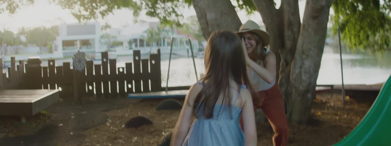A little girl slides down a slide in a park as her mother stands near a tree with a video recorder and smiles as she follows the girl with her camera