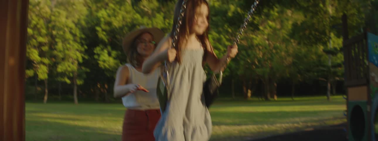 A woman wearing a sunhat and glasses is pushing her daughter on a swing at the park