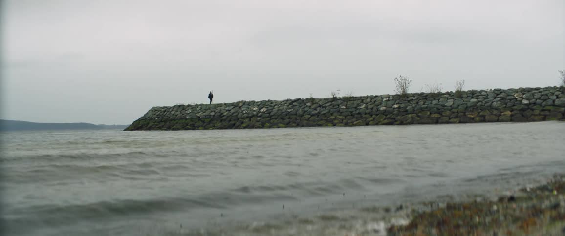 Person walks along low promontory as waves crash on beach under overcast sky