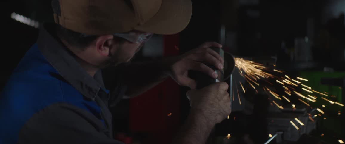 A man is polishing a metal with a tool while wearing cap and safety glasses