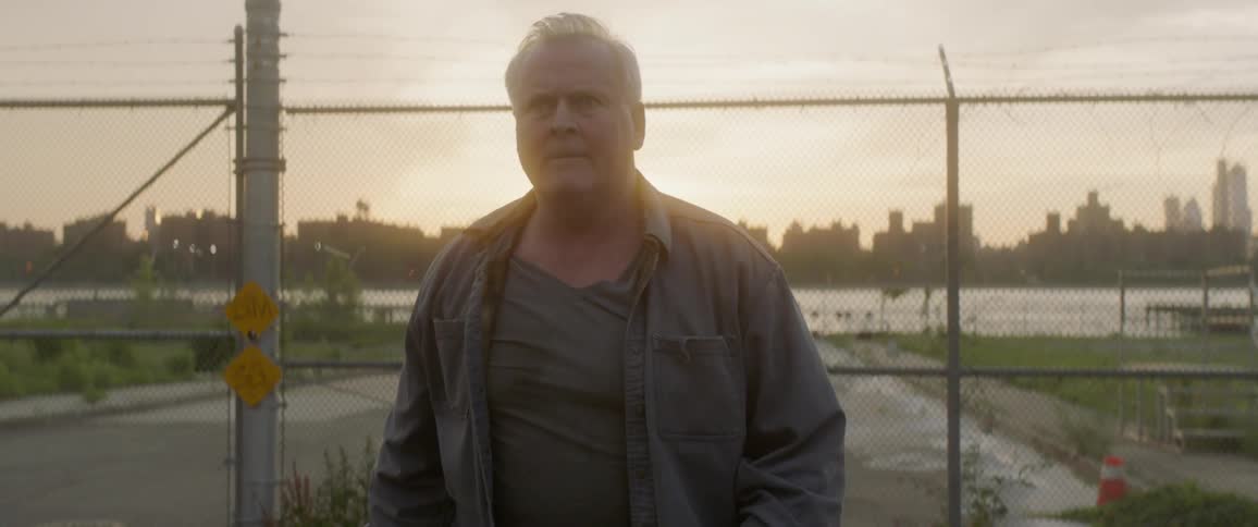 Older man walks aways from a chain link fence that's got a river and cityscape behind it as the sun sets