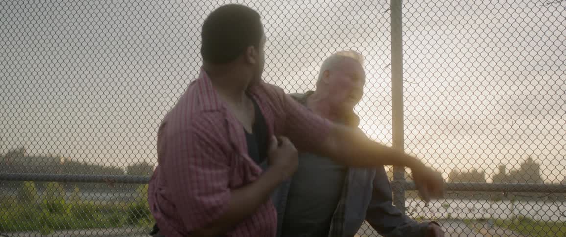 Two men viciously pummel each other by a fence in the light of a setting sun