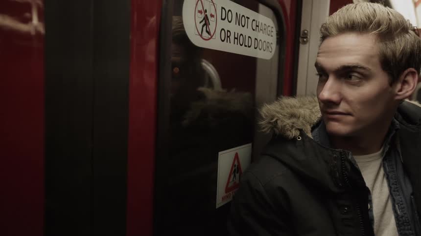 A young man stands on a train and looks out the window