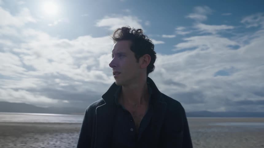 Man standing near coast with mountains in the background looks around on sunny day
