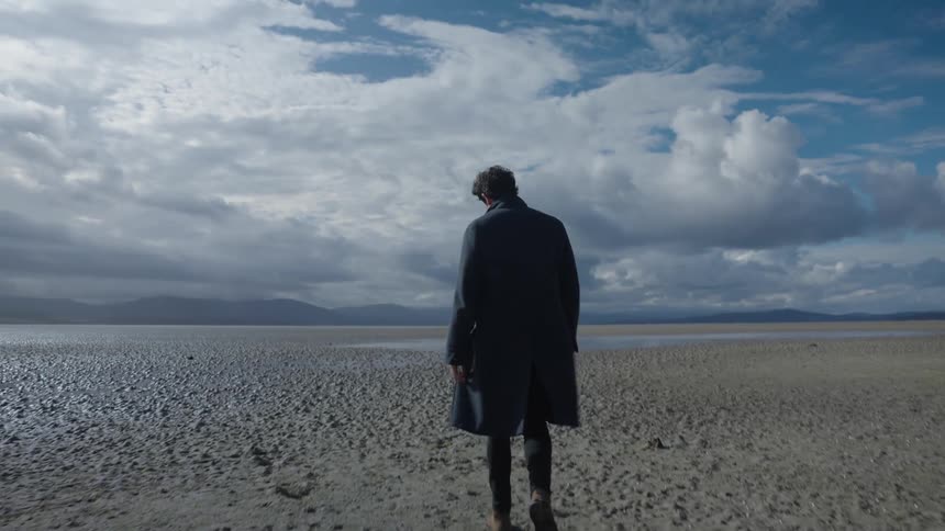 A man wearing an overcoat strolling across an open desert field during the day