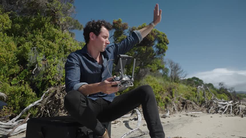 Man sits on case on beach using drone controller before making hand signals